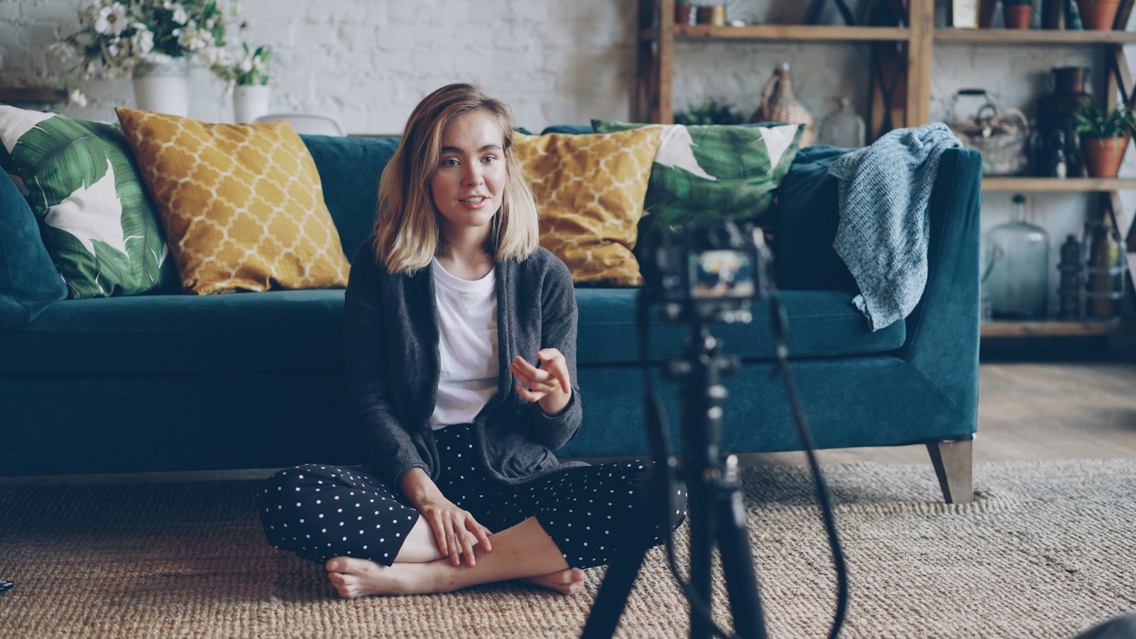 A course creator sitting on a living-room floor, recording a lesson on a camera on a tripod
